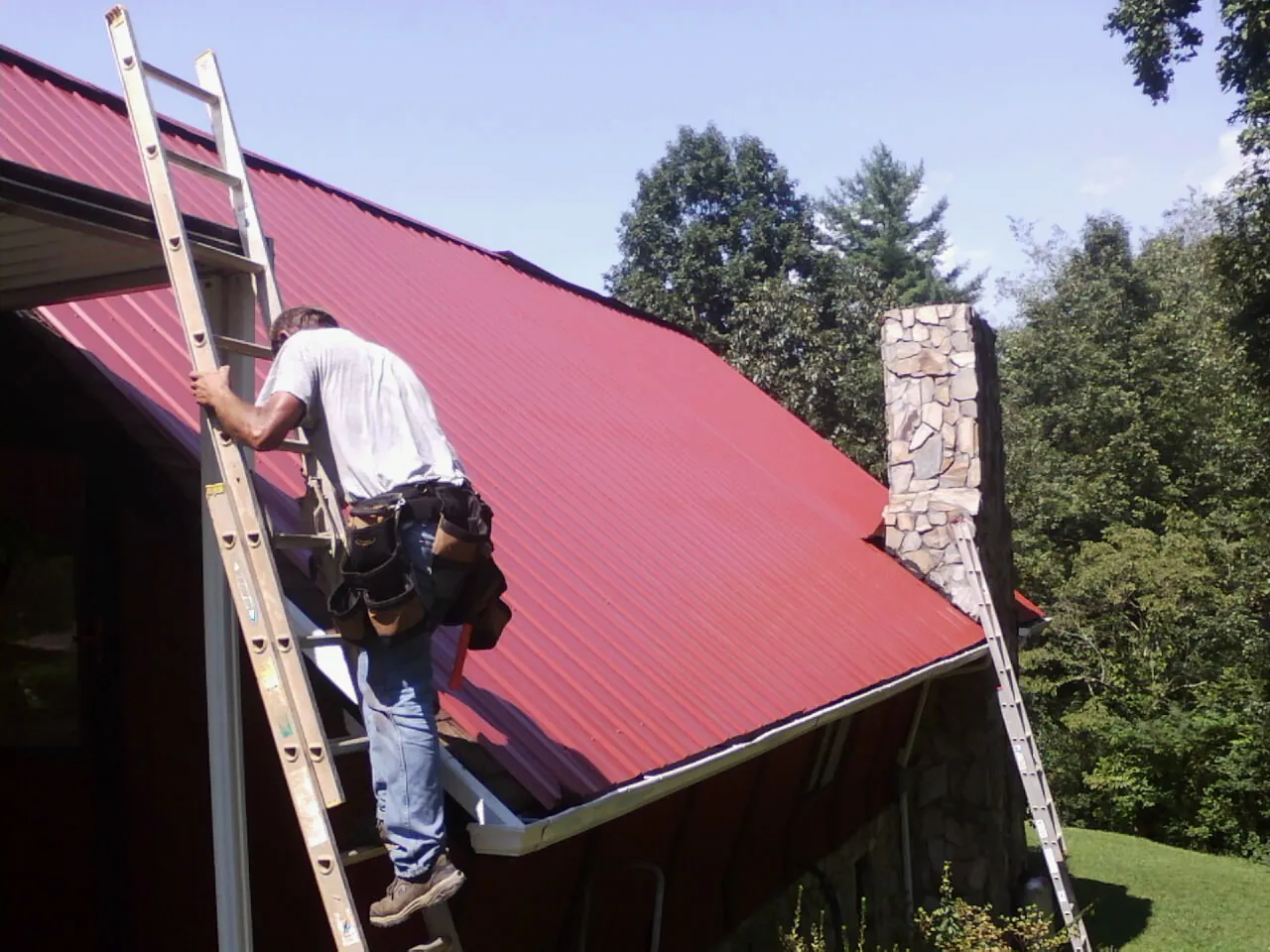 Roofer on a ladder beside a red metal roof and stone chimney — Soffit & Fascia Repair by Roofing Brothers in Diagonal